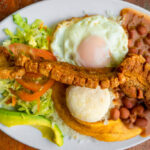 A "Bandeja Paisa" platter on a wooden table, featuring white rice, red beans, chicharrón (fried pork belly), ground beef, avocado, fried egg, plantains, arepas, and lemon wedge.