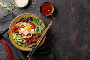 A bowl of Korean Bibimbap, a mixed rice dish, with carrots, spinach, mushrooms, bean sprouts, a fried egg, sesame seeds, nori strips, and gochujang sauce.